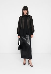 Donna che indossa una blusa nera trasparente a maniche lunghe, una gonna midi in pelle nera, sandali neri con tacco alto e tiene in mano una borsa nera.