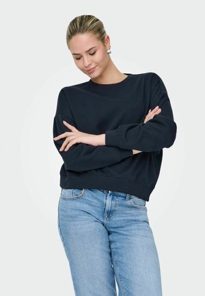 Sweat-shirt noir à manches longues et col rond, possédant une coupe décontractée. Associé à un jean en denim bleu clair, avec des détails minimalistes.