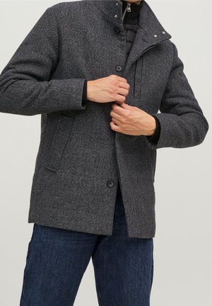 Personne boutonnant un manteau texturé gris foncé par-dessus une veste noire à fermeture éclair, portant un jean bleu, sur un fond clair uni.