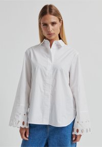 Chemise blanche à boutons avec manches longues, présentant des découpes circulaires au niveau des poignets. Fabriquée en tissu lisse et léger. Col classique.