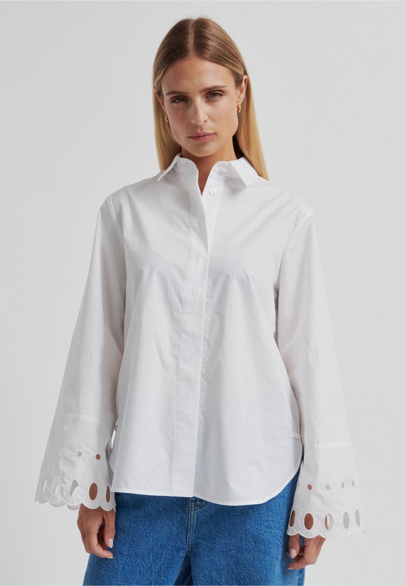 Chemise blanche à boutons avec manches longues, présentant des découpes circulaires au niveau des poignets. Fabriquée en tissu lisse et léger. Col classique.