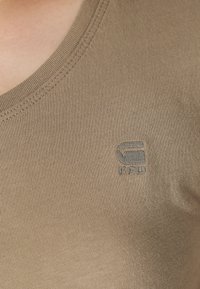 Šviesiai rudas V formos marškinėliai pagaminti iš lygaus, elastingo audinio, su iškaltu logotipu kairėje krūtinės pusėje, pabrėžiant minimalizmo dizaino elementus.