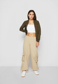 Oliven grønn bomberjakke, hvit crop top, beige cargo bukser med sidelommer, og hvite sneakers med oransje aksenter. Glatte stoffteksturer.