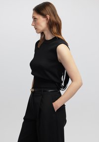 Maglione senza maniche nero con collo alto e tessuto testurizzato, abbinato a pantaloni neri a vita alta con una cintura nera elegante e dettagli dorati.