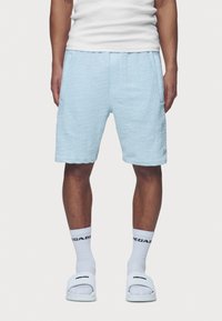 Shorts bleu clair texturés avec une ceinture élastique, dotés de poches latérales et d'un petit écusson logo. Portés avec des chaussettes blanches et des sandales.