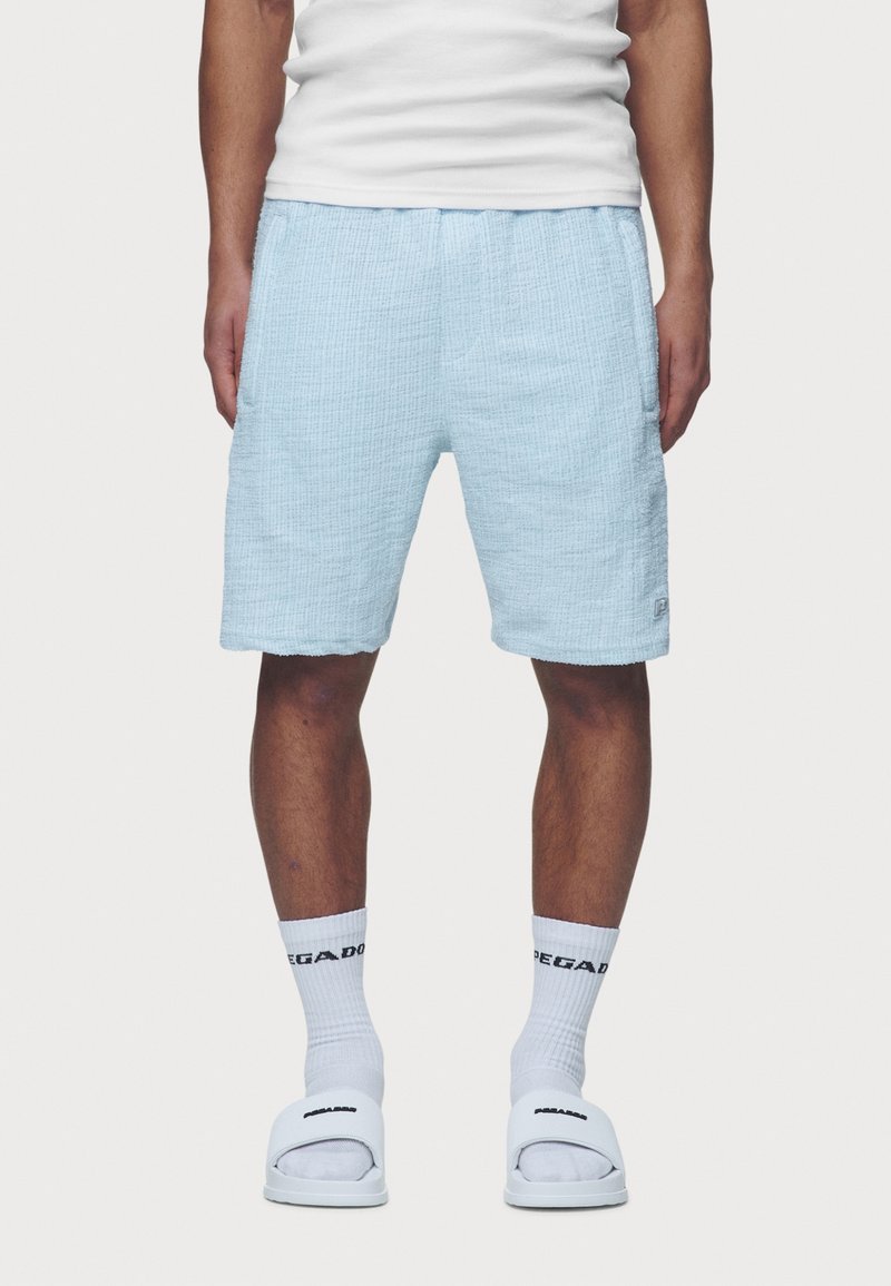Shorts bleu clair texturés avec une ceinture élastique, dotés de poches latérales et d'un petit écusson logo. Portés avec des chaussettes blanches et des sandales.