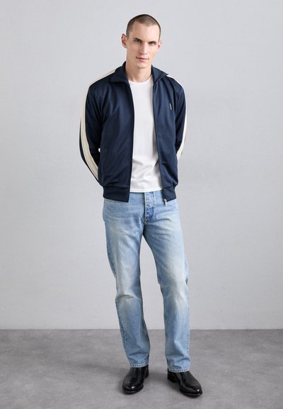 Marinblå zip-up jacka med krämfärgade ränder, vit t-shirt, ljusblå jeans och svarta skor. Jackan har en ståkrage och sidofickor.