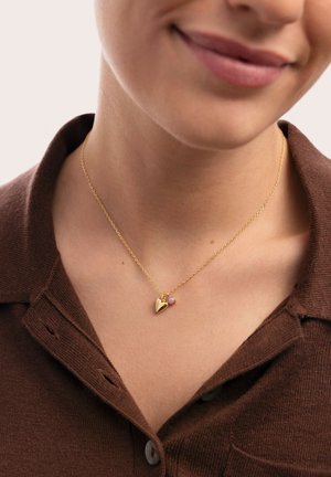 Donna che indossa una collana d'oro con un ciondolo a forma di cuore e una piccola perla rosa, vestita con una camicia marrone con colletto.