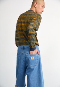 Long-sleeve top in dark green with orange wavy stripes, paired with light blue denim shorts featuring a back pocket logo patch.