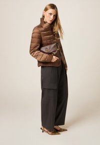 Braune Pufferjacke mit hohem Kragen, kombiniert mit weit geschnittenen dunklen Hosen. Die Accessoires bestehen aus einer strukturierten braunen Clutch und Schuhen mit Tierdruck.