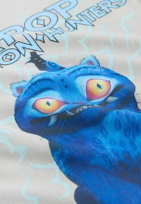 Blaues grafisches T-Shirt mit einer Cartoon-Katze mit übertriebenen Merkmalen und großen Augen. Der Text lautet "POP ON HUNTERS" vor hellem Hintergrund.