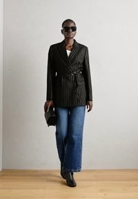 Svart blazer med pinstriper og belte, båret over en hvit topp og blå, vide jeans. Modellen holder en svart clutch og har på seg oversized solbriller.