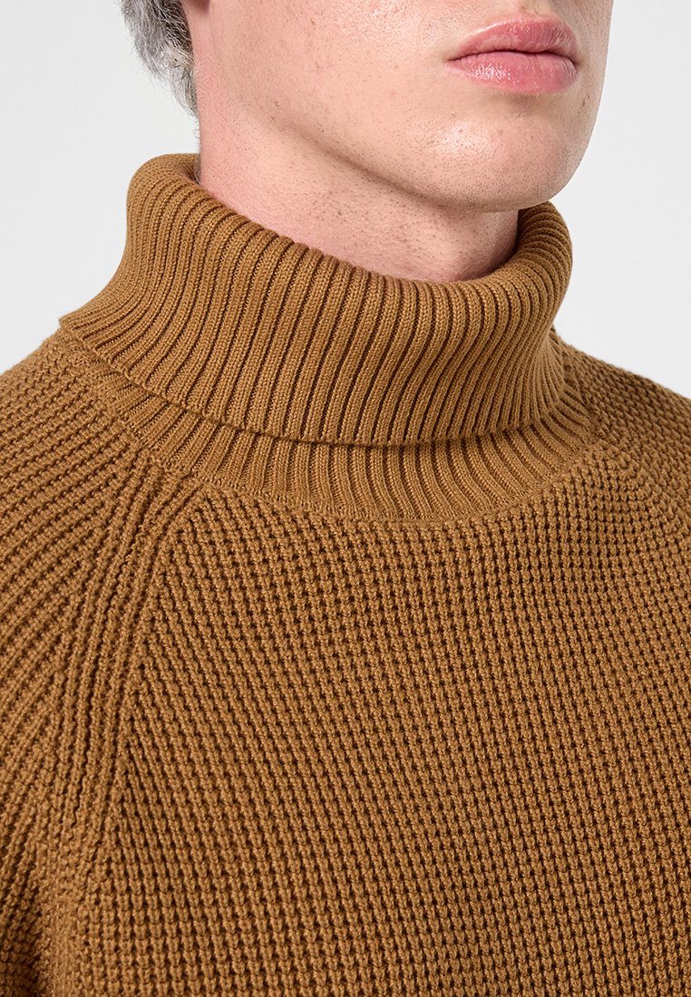 Maglione a collo alto lavorato a maglia in marrone, con una texture a coste, colletto alto e maniche raglan. Primo piano sulla zona del collo.