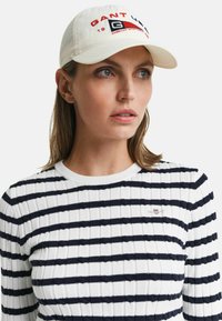 Casquette de baseball blanche avec des accents rouges et bleu marine, mettant en avant le logo "GANT". Le modèle porte un pull côtelé blanc et bleu marine à rayures avec un col rond.