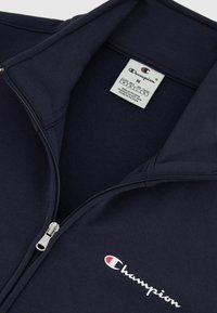 Chaqueta de forro polar azul marino con cremallera, que presenta un cuello alto, cremallera plateada y el logo bordado de Champion en blanco con acentos rojos.