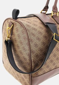 Sac de voyage à motifs marron avec des accessoires en laiton doré, présentant une bandoulière en tissu noir et des accents en cuir. Fermeture zippée en haut.