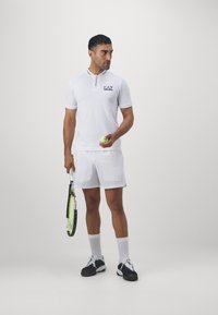 Polo deportivo blanco con cremallera y detalles en negro; combinado con pantalones cortos blancos. Sosteniendo una raqueta de tenis y una pelota; vistiendo zapatillas negras.