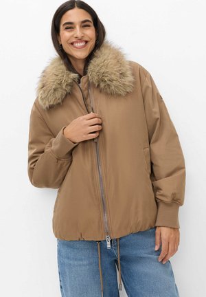 Smilende kvinde iført en beige dunjakke med en stor pelskrave af imiteret pels og blå jeans, poserer mod en hvid baggrund.