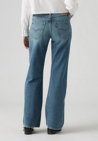 Jean flare en denim bleu avec taille haute, couture visible, deux poches arrière et une finition délavée, associés à des chaussures noires.