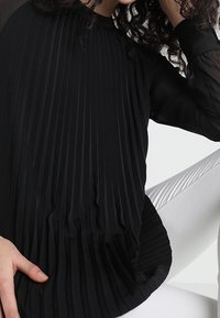Blusa nera plissettata con collo alto e maniche lunghe trasparenti, caratterizzata da pieghe verticali sul davanti e un pannello posteriore liscio.