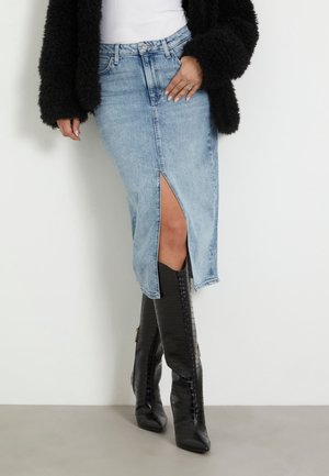 Midilengte denim rok in lichtblauw met een voorste split, gecombineerd met zwarte kniehoge laarzen met krokodillenprint. Gestructureerd zwart jasje.