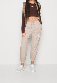 Beige joggers med elastisk midja och vitt Nike-logga, ihop med en brun långärmad crop top och en brun pufferväst. Grå sneakers.
