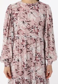 Robe rose clair avec un motif floral dans des nuances de rose et de crème, manches bouffantes longues, encolure ronde et ourlet à volants en couches.