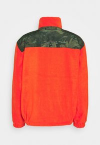 Oranžová fleecová bunda s vysokým límcem, zadní část s maskáčovým vzorem a elastické manžety pro těsné přilnutí.