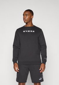 Black sweatshirt with "HYROX" logo in white, round neck, long sleeves. Paired with black shorts featuring a subtle Puma logo. Smooth texture.