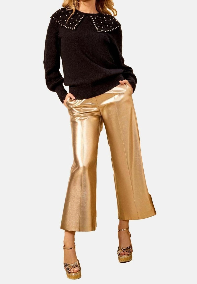 Femme portant un pull noir avec un col orné de perles, un pantalon large doré brillant et des talons à plateforme, debout les mains dans les poches.