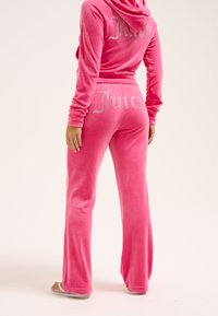 Roza velour trenirka s kapuco, z logotipom "Juicy" iz kristalov na hrbtu zgornjega dela in na hlačah. Hlače so razširjene, rokavi pa dolgi.