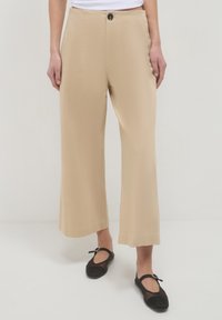 Pantaloni a culisse beige a gamba larga indossati con scarpe Mary Jane in rete nere su uno sfondo neutro.