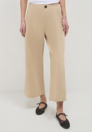 Beige, weit geschnittene, knielange Hose, getragen mit schwarzen Netz-Mary-Jane-Ballerinas vor einem neutralen Hintergrund.