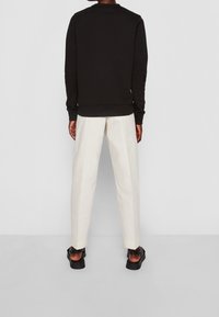 Zwarte sweatshirt met ronde hals en geribbelde manchetten, gecombineerd met lichtgekleurde geplooide broeken en zwarte sandalen op een neutrale achtergrond.