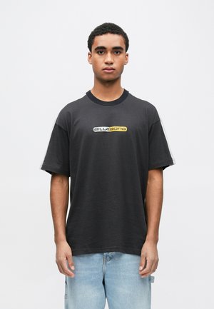 Mladić stoji okrenut prema naprijed, nosi crnu Billabong majicu i svijetloplave traperice, na jednostavnoj bijeloj pozadini.