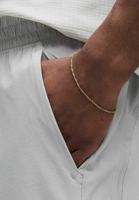 Gul kedjebracelet på ett handled, kombinerat med ljusgrå sportshorts. Tyget har en slät yta och elastisk midja.