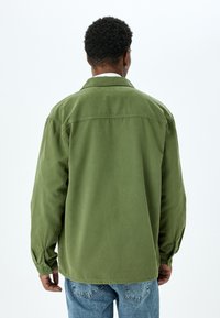 Mies, jolla on lyhyet kiharat hiukset, pukeutuneena oliivinvihreään pitkähihaiseen paitaan ja sinisiin farkkuihin, seisoo selin yksinkertaista taustaa vasten.