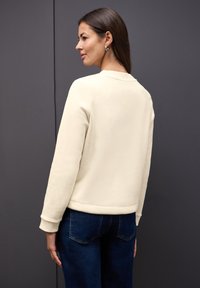 Sweatshirt de couleur crème avec un col rond et des poignets côtelés, associé à un jean bleu foncé. Texture lisse, design ajusté.