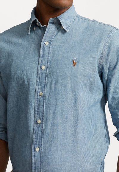 Light blue, button-up shirt made of denim fabric. Features a collar, white buttons, and an embroidered logo on the left chest.