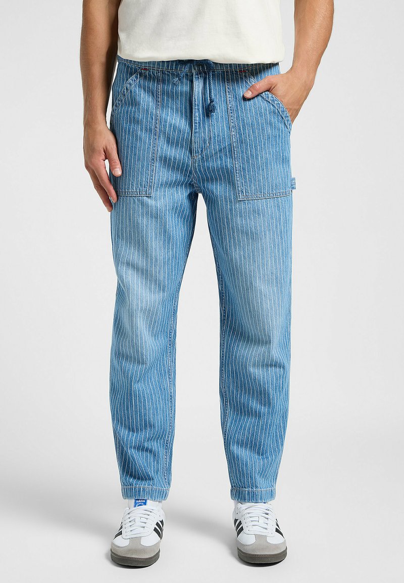 Pantaloni in denim a righe azzurro chiaro con una vestibilità rilassata, caratterizzati da una cintura elastica e tasche frontali. Indossati con sneakers grigie e bianche.