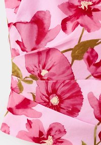 Tissu avec un grand imprimé floral rose et rouge avec des tiges et feuilles vertes sur fond rose clair, montrant un détail de couture pliée.
