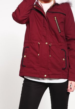 Vrouw die een rode winterjas met rits draagt, voorzien van bontkraag, gouden knopen en zwarte broek, staand met één hand bij haar hoofd.