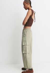 Top scurt maro, texturat, cu spatele deschis; pantaloni cargo lejeri, verzi deschis, cu buzunare laterale; pantofi maro cu sireturi crem.