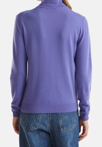 Fioletowy sweter z długim rękawem i wysokim kołnierzem, z prążkowanymi mankietami, noszony na niebieskich dżinsach. Miękka faktura, prosty design.