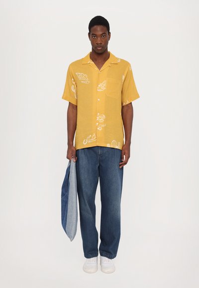 Kortermet gul skjorte med hvite mønstre, kombinert med blå denimbukser. Personen holder en sammenfoldet denimjakke. Hvite joggesko.