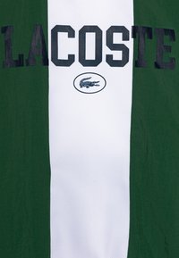 Tessuto a righe verdi e bianche con "LACOSTE" stampato in grassetto lettere nere e un logo di coccodrillo nero all'interno di un ovale bianco. Materiale testurizzato.