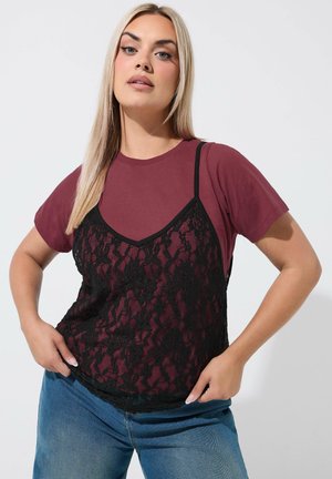 Blondynka ubrana w bordową koszulkę pod czarną koronkową koszulką, niebieskie dżinsy, ręce na biodrach, lekki makijaż, stoi na białym tle.
