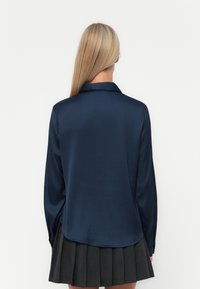 Navy satin shirt with a collar, long sleeves, and a curved hem. Textured fabric with a smooth finish, paired with a pleated skirt.