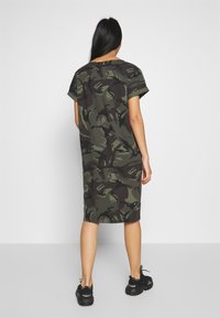 Robe à manches courtes, longueur genou, en imprimé camouflage vert foncé, fabriquée en tissu doux. Présente une coupe décontractée et un col ras du cou. Portée avec des baskets noires.