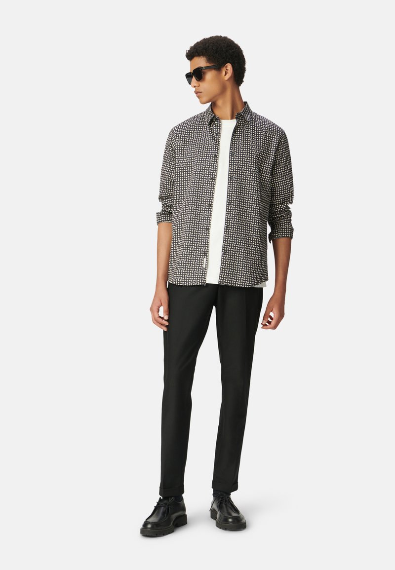 Camicia a fantasia bianca e nera, in materiale leggero, con design a bottoni, abbinata a pantaloni neri e scarpe nere. Gli accessori includono occhiali da sole.